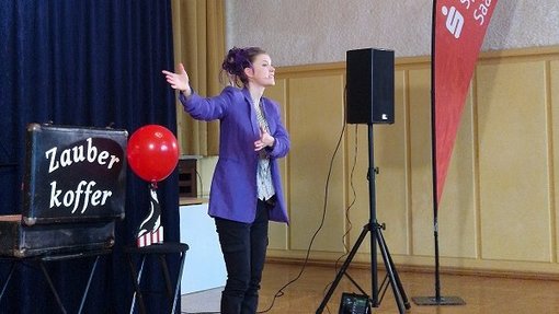 Zauberin mit lila Jacke auf der Bühne mit dem Zauberkoffer und einem roten Luftballon